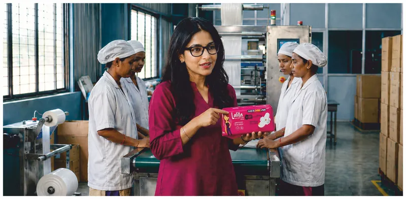 A woman holding a pack of Lalita Sanitary Pads on the factory floor, flanked by two factory workers in white uniforms.